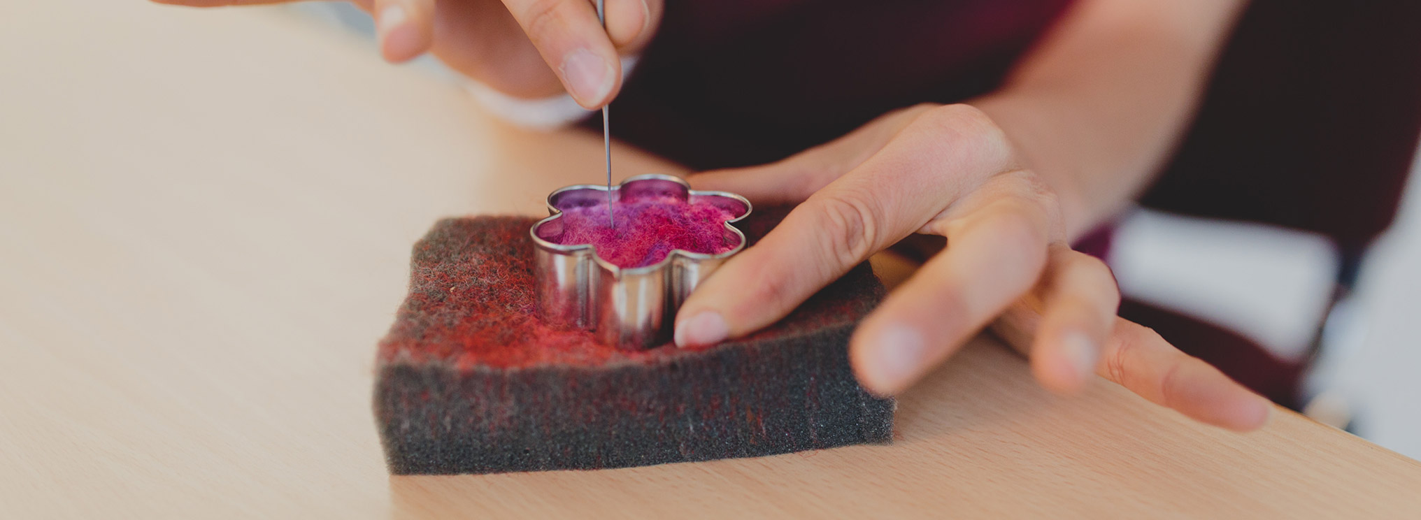 Eine Hand erstellt mit einer Filznadel eine Blume aus Wolle in einer Keksform.