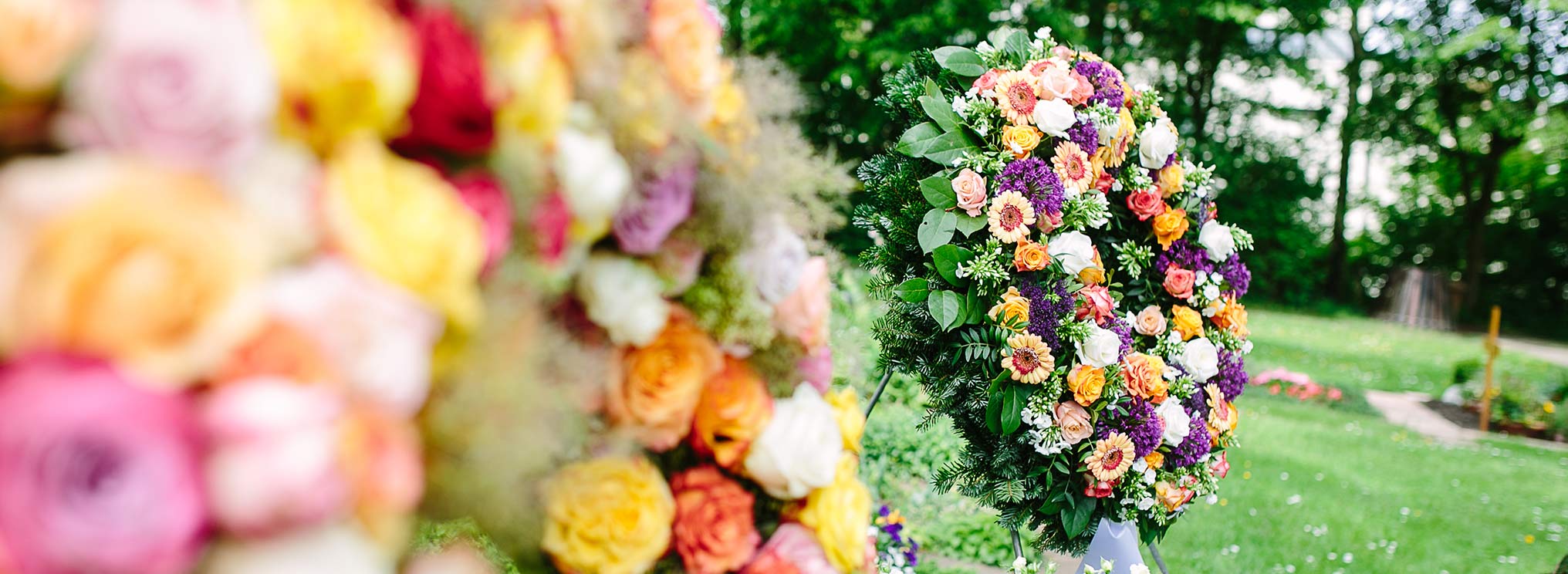 Nahaufnahme eines Blumengestecks mit Rosen, umgeben von weiteren farbenfrohen Blumenkränzen auf einer grünen Wiese.